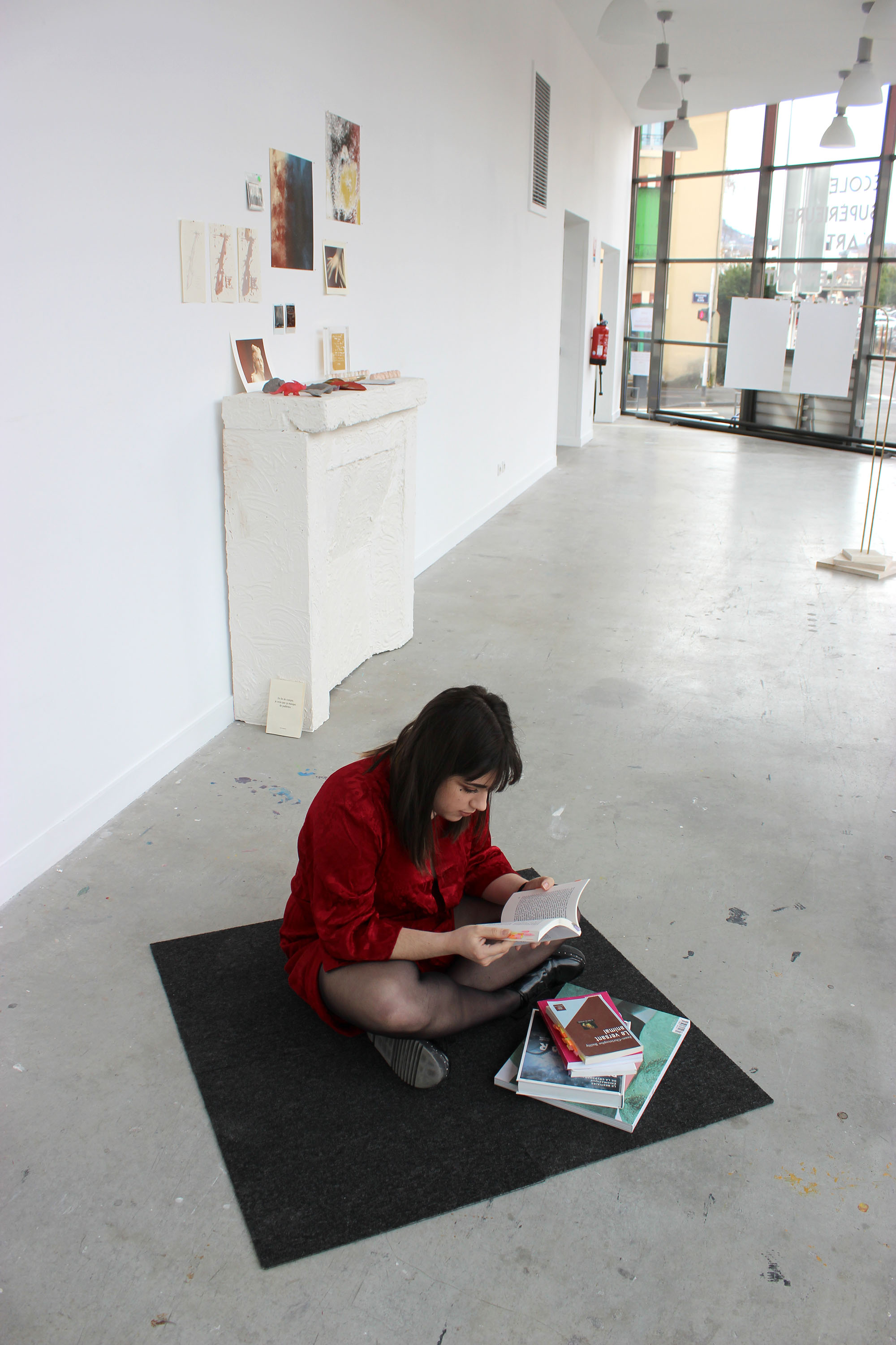 Dans un couloir, une jeune fille lit assise sur un carré de moquette. Une pile de livres est posée à côté d'elle. Les livres et la jeune fille ne sont pas les mêmes que précédemment.